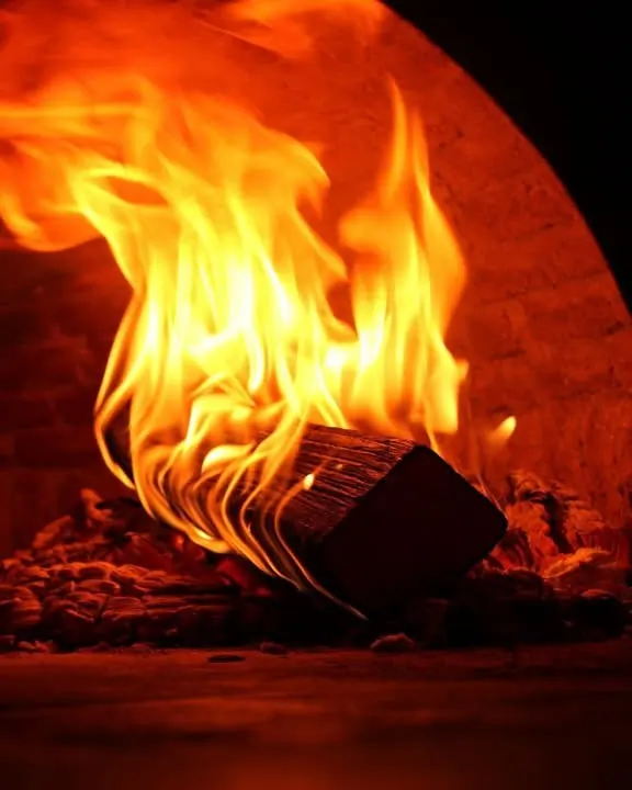 Bûche en feu sur le lit de braises dans le four à pizza d'Il Piacere, flammes orange éclatant sur fond noir
