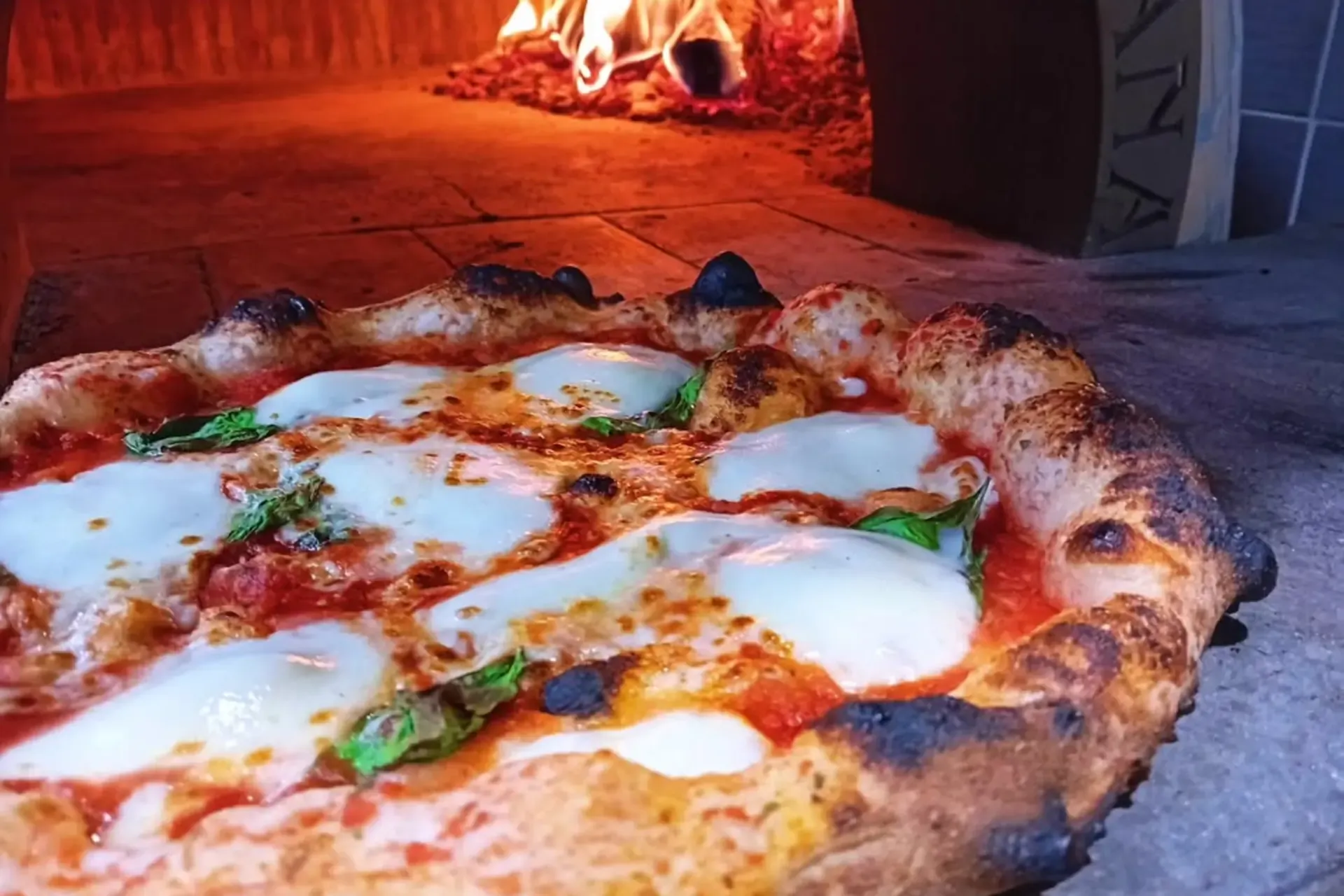 Pizza Margherita sortant du four à bois d'Il Piacere, mozzarella bouillonnée, bords du cornicione noircis, flammes visibles à l'arrière dans la gueule du four