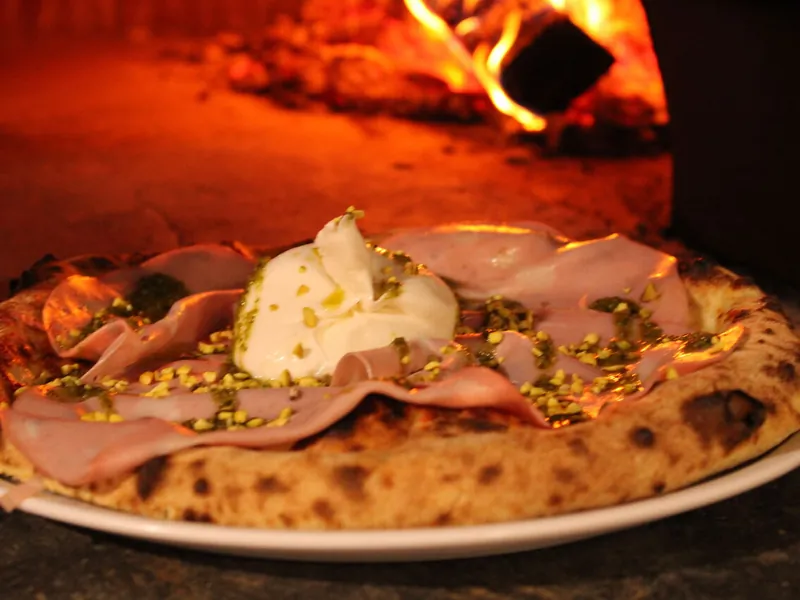 Pizza Mortadellina d'Il Piacere : base mozzarella, mortadelle, burrata centrale et pistaches concassées, flammes du four en arrière-plan flou
