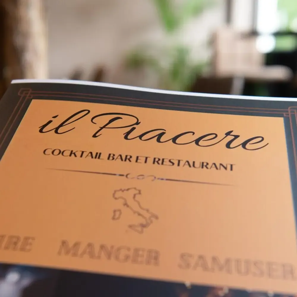 Carte papier d'Il Piacere posée sur une table, titre Cocktail Bar et Restaurant avec silhouette de l'Italie, mention Boire Manger S'amuser
